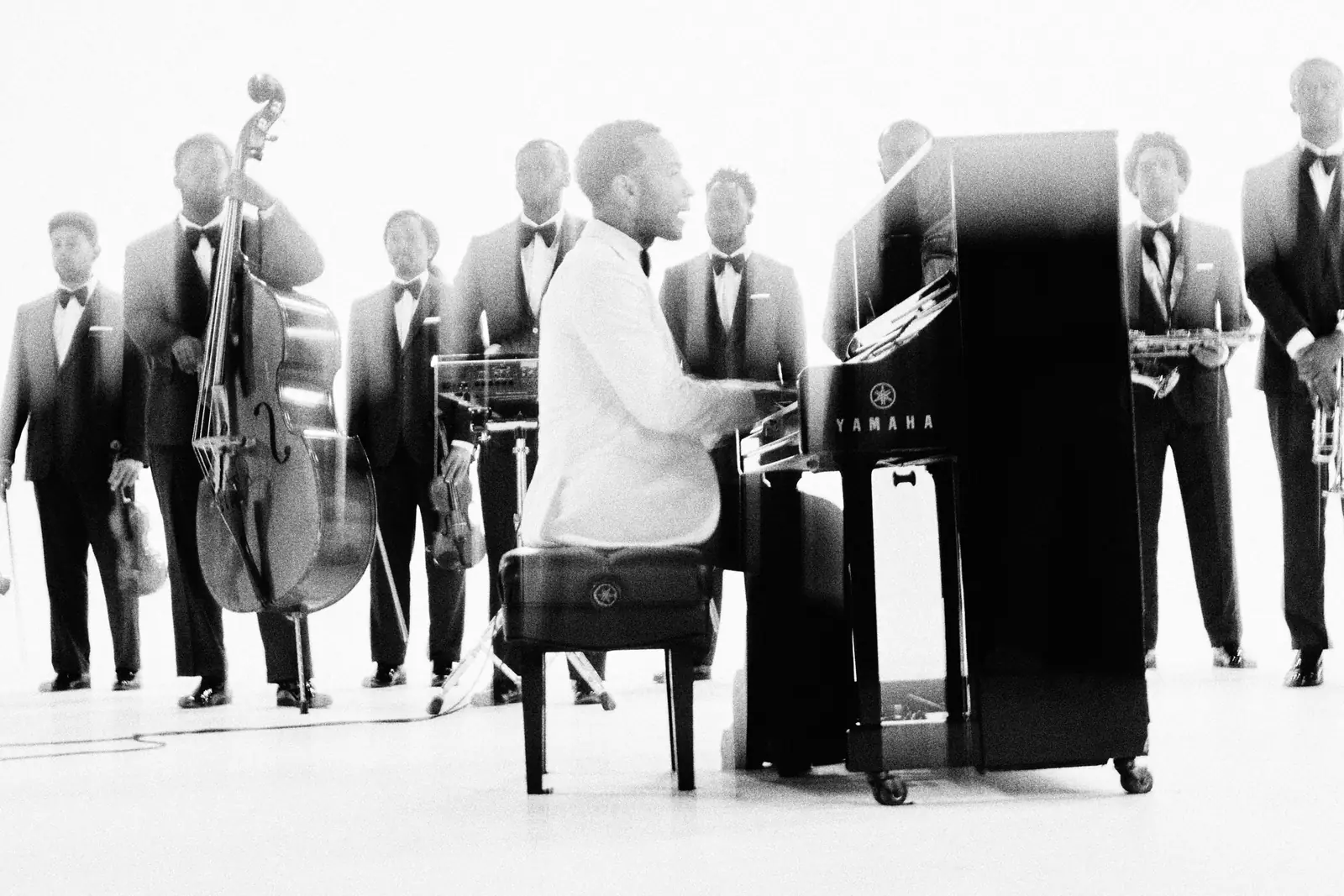 John Legend at the piano in a white studio, a big band in black tuxedos standing behind him
