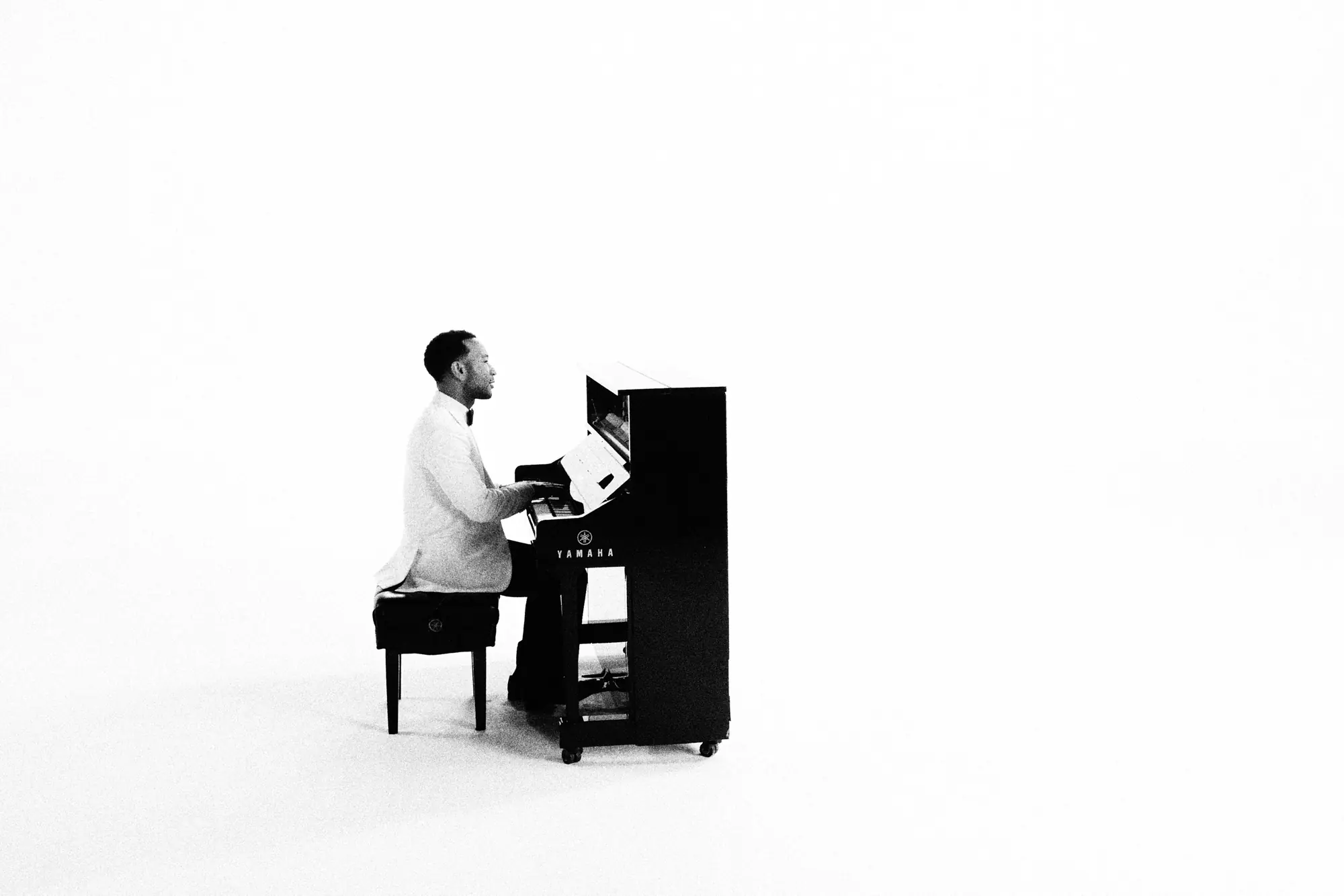 John Legend in a white dinner jacket at an upright piano in an empty white studio